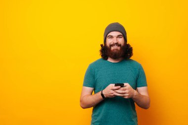 Güzel telefon tutarak ve kameraya boşaltmak ile sarı arka plan üzerinde seyir t-shirt erkekte hipster sakallı