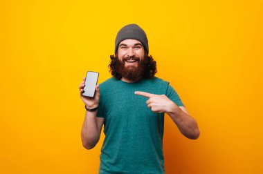 Neşeli sakallı hippi erkek gülümseyen ve smartphone sarı arka plan üzerinde poiting