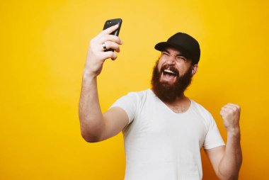 Cep telefonu görünümlü ve sarı arka plan üzerinde izole kutlama beyaz t-shirt mutlu bir genç sakallı adam portresi