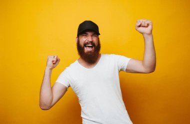İzole sarı arka plan üzerinde beyaz t-shirt kutluyor başarı memnun Genç sakallı adam portresi