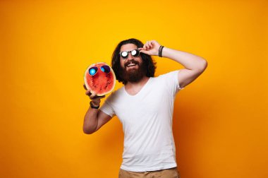 Half of Watermelon face with glasses held by a bearded man. Hispter wearing white shirt with long hair. Summer offer, vacation, holiday rest. Traveling and tourism concept. Summer buddy. Vegetarian or Vegan concept. clipart