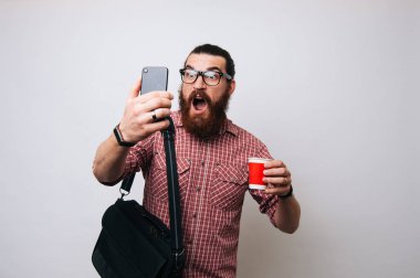 Çalışmaya, onun smartphone gidiş arıyorsunuz şaşırmış sakallı adam. Telefonun başında online para kazanmak. Kırmızı ekose gömlek kırmızı kağıt bardak çay ya da kahve, cappucino veya latte ile tutarak çalışma çantalı adam.