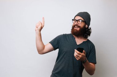 Modern Yenilikçi genç ve sakallı adam bir teklif ya da bir anlaşma işaret ediyor. Adam Gri tişört ve kürk şapka şapka ve onun radyo earphones earpods giyer. Adam holding smartphone. En iyi teklif kavramı.
