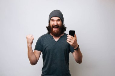 Neşeli genç gri T-shirt giyen adam sakallı ve kürk şapka şapka ve mutlu. Kazanan adam kazanan jest beyaz arka plan üzerinde gösterilen smartphone ile.
