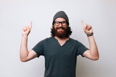 Neşeli genç bir T-shirt ve gözlük ve bir kürk şapka kap giyen adam sakallı. Elleri ve yukarı işaret parmakları ile işaret eden adam. Teklif kavramı.