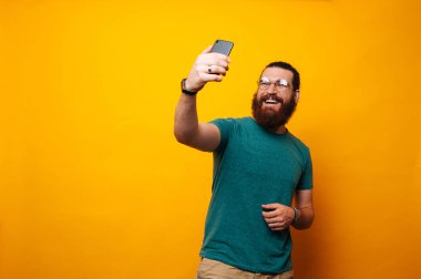 Mutlu gülümseyerek gözlük takıyor ve selfie ile smartphone üzerinde sarı arka plana alarak hipster adam sakallı