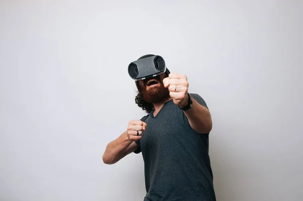 Hipster with excited face fighting box in virtual reality with modern digital gadget. Man with beard in VR glasses boxing, white background. Virtual sport boxing lessons concept.