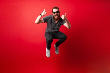 Cheerful bearded hipster man with sunglasses jump over red background and showing thumbs up clipart