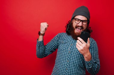 Zaferini kutluyor ve smartphone üzerinde kırmızı arka plan kullanarak neşeli sakallı adam portresi