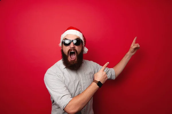 カジュアルな身に着けているサンタ クロース帽子さして提供のため Copyspace で赤い背景の上のひげを生やした男を驚かせた — ストック写真