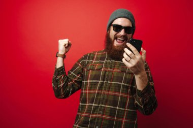 Mutlu neşeli başarı kutluyor ve smartphone kırmızı arka plan üzerinde tutan adam sakallı
