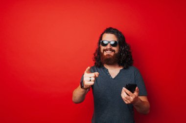 Akıllı telefon tutarak ve kameraya kırmızı arka plan üzerinde işaret mutlu ve neşeli sakallı adam