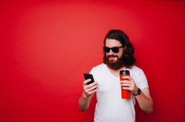 Sakallı adam akıllı telefon ve içki kahve kırmızı arka plan üzerinde kullanma