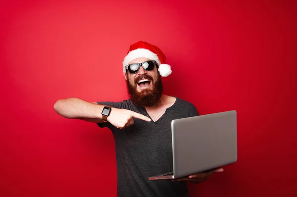 サンタ クロースの帽子の赤い背景の上のノート パソコンを指すと陽気なひげを生やした男 — ストック写真