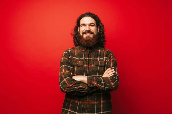 赤い背景の上の交差の腕を持つ陽気なひげを生やした流行に敏感な男性の肖像画 — ストック写真