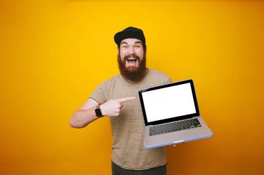Amazed man with beard pointing at blank screen on laptop over yellow background clipart