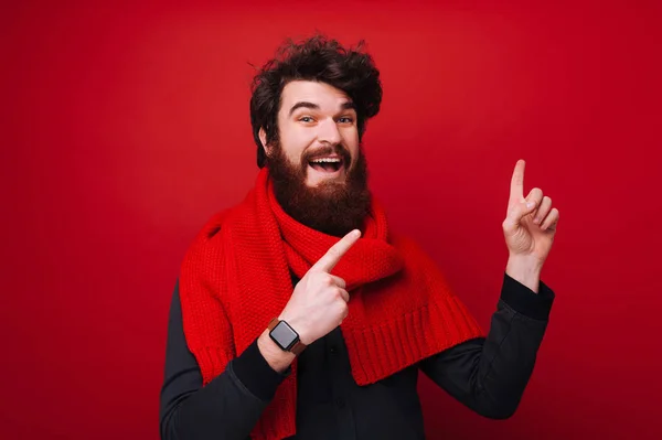 Portrait of cheerful bearded hipster man pointing away over red background at copyspace
