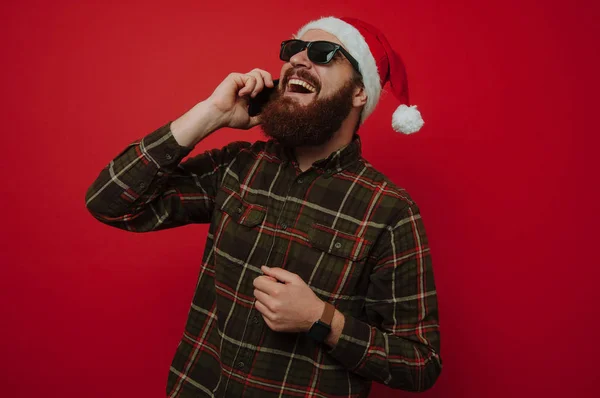 Happy smiling bearded hipster man with santa claus hat talking on phone over red background