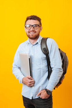Genç öğrenci holding dizüstü gülümseyen ve sırt çantası sarı arka plan üzerinde giyen portre