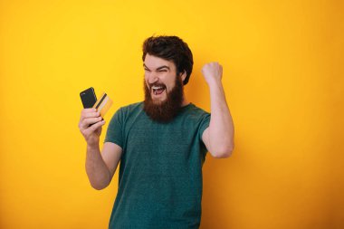 Cep telefonu ve kredi kartı izole sarı artalan üzerinde tutarak tshirt heyecanlı bir sakallı adam portresi kutluyor 