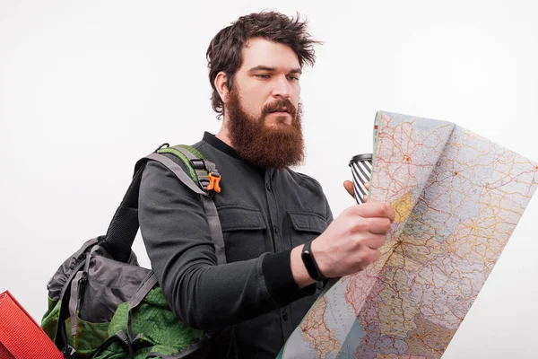 Male tourist enjoying travel  with paper map in hands looking for destination, trendy hipster guy holding takeaway coffee and thinking about next showplace