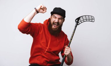 Floorball concept, exited bearded man in red hoody holding stick and doing win gesture with rised hand over white background  clipart