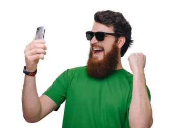 Heyecanlı genç sakallı adam ayakta cep telefonuyla wihite duvar arka plan üzerinde izole ve kazanan bir jest yapar