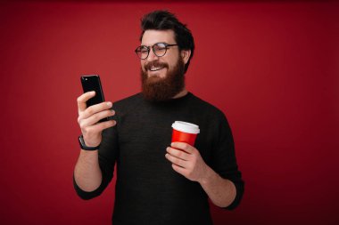 Mutlu gülümseyerek telefon ekranına bakarak ve mektuplarını kontrol ederken kahve tutan adam sakallı