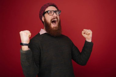 Şanslı genç hipster adam kırmızı arka plan üzerinde izole. Sakallı adam gözlük ve şapka giyen bir şey kazanmış gibi davranır