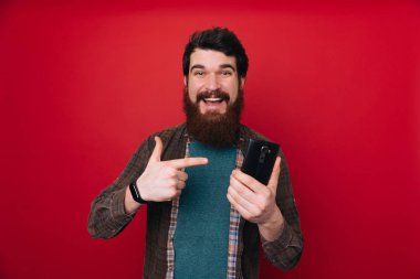 Mutlu tutarak ve cep telefonu kırmızı arka plan üzerinde işaret ekose gömlekli sakallı genç adam 