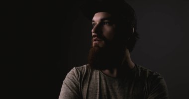 Sakallı yakışıklı tshirt ve çapraz kolları uzakta arka plan karanlık üzerinde seyir şapka