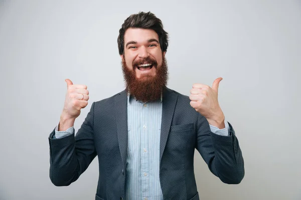 Portrait of excited man with opened mouth dressed in formal wear giving thumbs-up against gray background