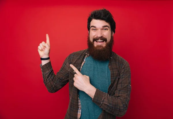 Handsome man with beard pointing with the index fingersup  and looking at camera on isolated red background