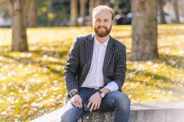 Genç adam, rahat kıyafetler giymiş kentsel arka planda kamera bakarak modeli sakallı. Checkerd takım elbiseli parapetin üzerinde oturan sakallı adam 