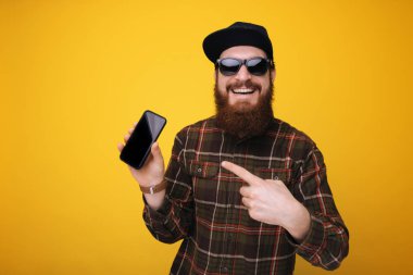 Onun smartphone vasıl sarı arka plan üzerinde işaret mutlu sakallı adamın şapka ve güneş gözlüğü, giydiğini