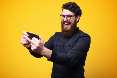 Heyecanlı genç sakallı adam sarı duvar arka plan üzerinde izole ayakta görüntüsünü oyun oyunlar cep telefonu tarafından.