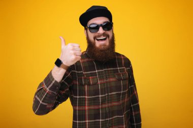Yakışıklı bir şapka ile adam ve sarı bir arka plan üzerinde Studio gösterir başparmak yukarı sakal. 