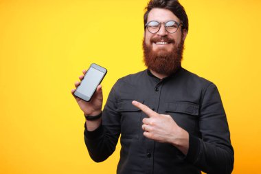 Görüntü esmer smartphone vasıl parmak işaret ve sarı arka plan üzerinde gülümseyen adam sakallı