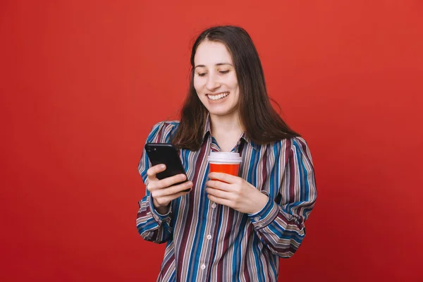 一个休闲的年轻女子的肖像孤立在红色背景 拿着外卖咖啡用手机 — 图库照片