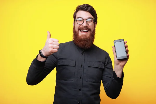 Jovem Barbudo Homem Segurando Smartphone Gesticulando Polegar Sobre Fundo Amarelo — Fotografia de Stock