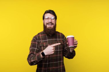 Mutlu gülümseyen genç adam kahverengi kareli gömlek poiting fincan kahve küreği çay, sakallı