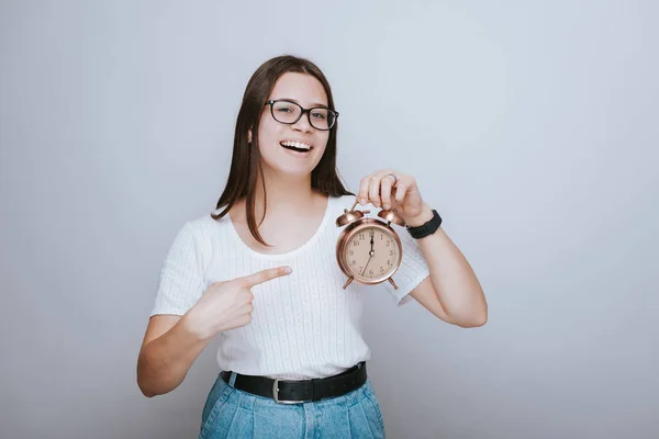 Joven chica alegre está sosteniendo un reloj despertador y apuntando a ella — Foto de Stock