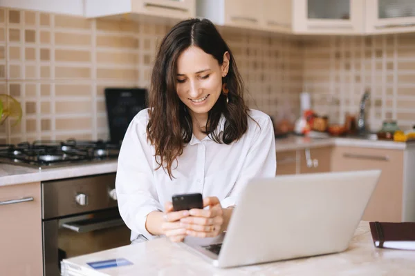 Krásná Usměvavá Žena Používající Telefon Při Práci Laptopu Kuchyňském Pokoji — Stock fotografie
