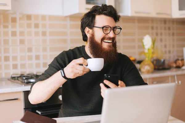 Joven Con Barba Escribiendo Portátil Está Sentado Cocina Sonriendo Debido —  Fotos de Stock