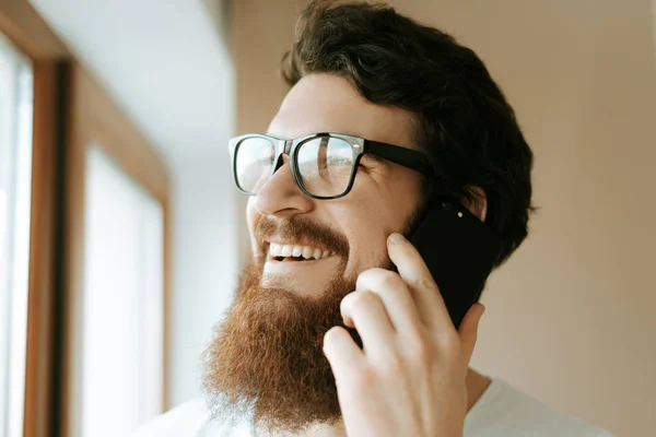 A photo of bearded man wearing glasses, is talking on mobile standing near window at home