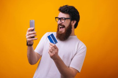 Sarı arka plan üzerinden online alışveriş için akıllı telefon ve kredi kartı kullanarak heyecanlı sakallı adam 