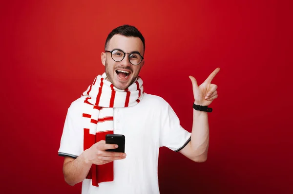 Handsome happy hipster with scarf and glasses holding mobile and pointing away over red background