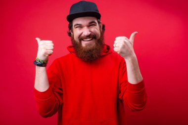 Neşeli yakışıklı sakallı adam kameraya bakıyor ve kırmızı arka plan duvar üzerinde başparmak gösteren