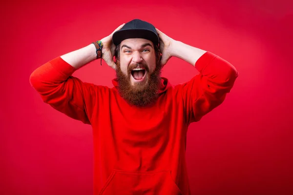 Handsome frustrated guy with beard screaming and putting hand on head, standing over red isolated  background