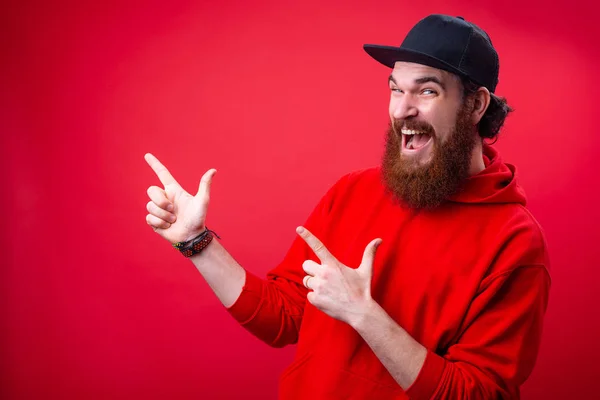 Excited bearded guy in red hoody and black hat, poitning with finger at copy space over red background